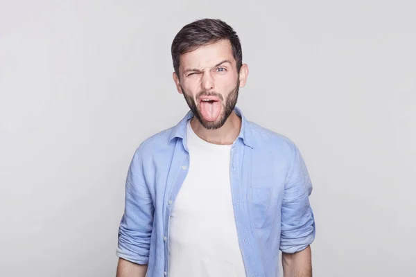 Emocional y barbudo joven guapo morena empleado con camisa azul mueca, haciendo bocas, sacando la lengua en la cámara tratando de burlarse de alguien, actuando como un niño travieso . — Foto de Stock