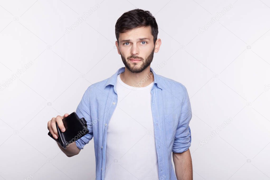 Young handsome Caucasian blue eyed handsome bearded man being confused and stressed about his empty wallet looking at camera with sad and disappointed face expression. Money, business, income concept.