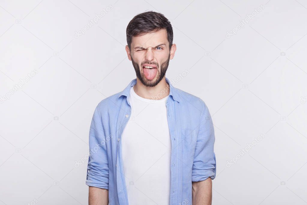 Displeased and bearded  young handsome blue eyed man employee wearing white shirt grimacing, making mouths, sticking out his tongue at camera trying to tease someone, acting like a little naughty child.