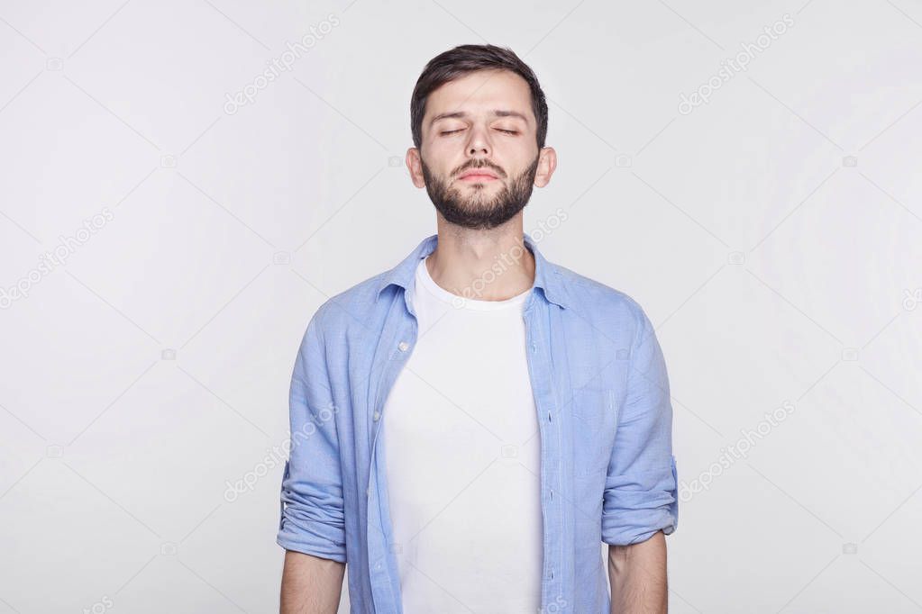 People and work. Exhausted and tired man with dark hair standing against white wall with eyes closed, thinking about something. Tired handsome Caucasian guy in blue jacket having rest at the office. 