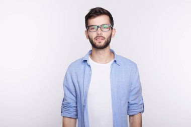 Duygu kendinden emin ve yakışıklı beyaz stüdyo duvar poz karşı beyaz genç hipster gözlüklü siyah çerçeve ve mavi gömlek kameraya kayıtsız taşlı yüz ifadesi ile seyir.