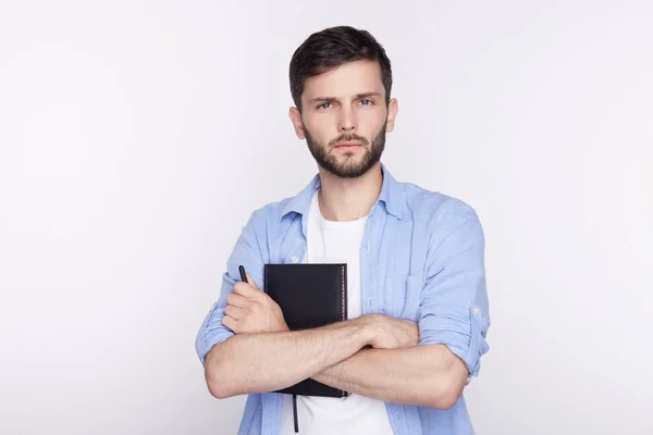 Imagen de un atractivo estudiante europeo serio con barba, bigote, vestido con una camiseta casual, mirando a la cámara con expresión facial pedregosa indiferente, manteniendo sus brazos cruzados en la pared blanca del estudio — Foto de Stock