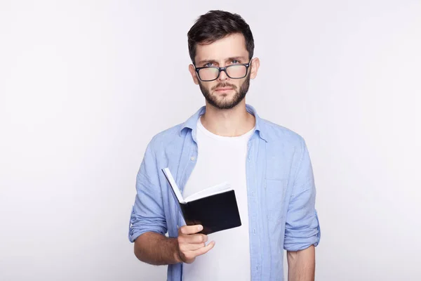 Casualmente vestido joven guapo estudiante barbudo con ojos azules con gafas se ha centrado mirada concentrada como lee la información en el libro de texto y tomar notas en copybook, preparándose para el examen . — Foto de Stock