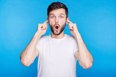 Genç adam rahat, giyinmiş dehşet içinde çığlık panik kameraya bakarak açık ağız. Kulakları, mavi duvar Gürültücü komşular ile kızgın kapsayan tahriş olmuş erkek bağırarak portresi kapatın.