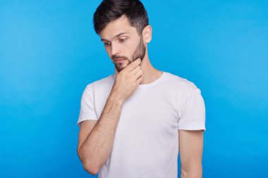 Çatık, sanki bir şey hatırlamaya çalışarak çenesini üzerinde el tutmak veya mavi duvar arka plan üzerinde karar düşünceli ifade, konsantre genç yakışıklı adam portresi stüdyo.