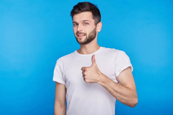 Estudio headshot de feliz joven caucásico hombre en camiseta blanca, haciendo gestos pulgares hacia arriba, diciéndole que usted está haciendo un buen trabajo, mirando y sonriendo a la cámara con expresión de confianza alegre . — Foto de Stock