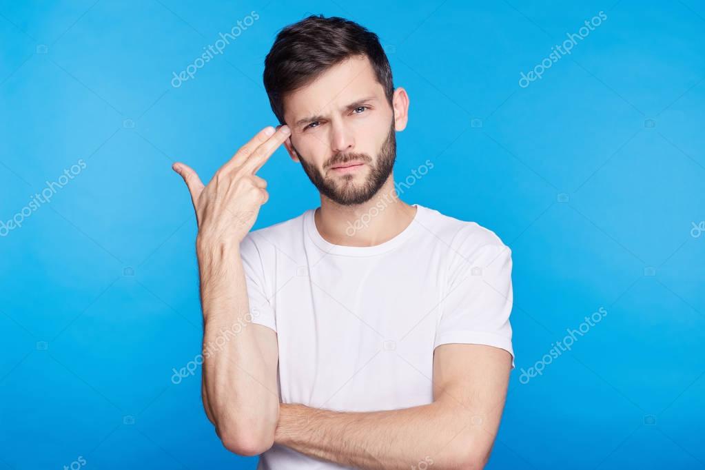 Close up isolated portrait of European young bearded hipster manager bored with work, shooting himself with finger gun gesture against blank blue wall background. Human face expressions and emotions.