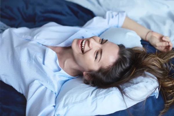 Attraente bruna allegra femminile che si estende in camera da letto mentre si trova a letto indossando camicia blu e jeans, ridendo ad alta voce con un sorriso affascinante, pieno di energia, sentendosi eccitato per il nuovo giorno felice . — Foto Stock