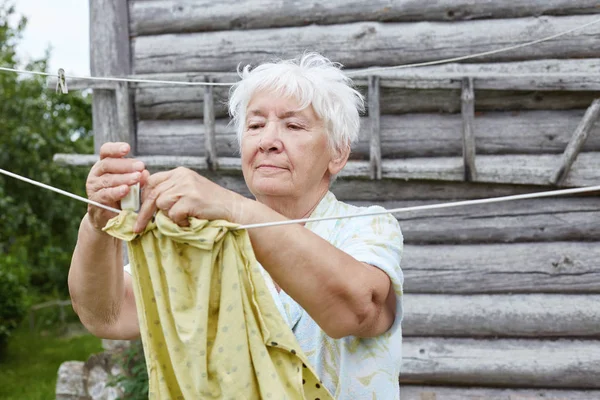 매력적인 회색 머리 백인 오래 된 회색 머리 여자의 본격적인 야외 초상화. 할머니는 목욕탕 배경 빨랫줄에 드라이로 셔츠를 걸려 있다. 마 생활, 농촌 현장. — 스톡 사진