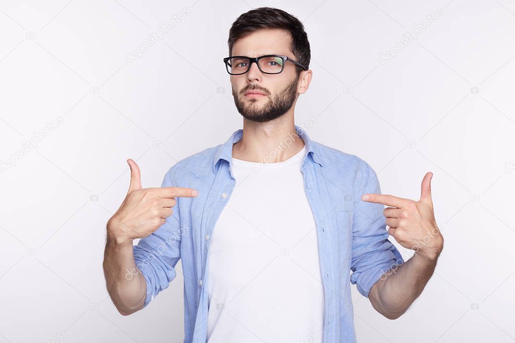 T-shirt design and advertising concept. Style and fashion. Indoor shot of cheerful smiling young Caucasian man with beard and blue eyes pointing index finger at copy space on his blank white t-shirt.