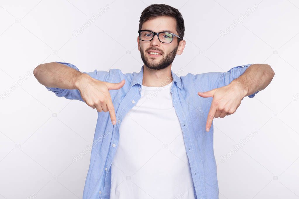 Unshaven stylish guy wearing white casual T-shirt and eyeglasses pointing at copy space for your print or advertisement indoors against studio white concrete wall. Design and advertising concept.