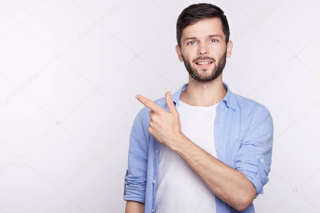 Horizontal portrait of young handsome brunette man with smile wearing casual blue shirt posing against white background pointing with index finger at white copy space for your advertisement content.