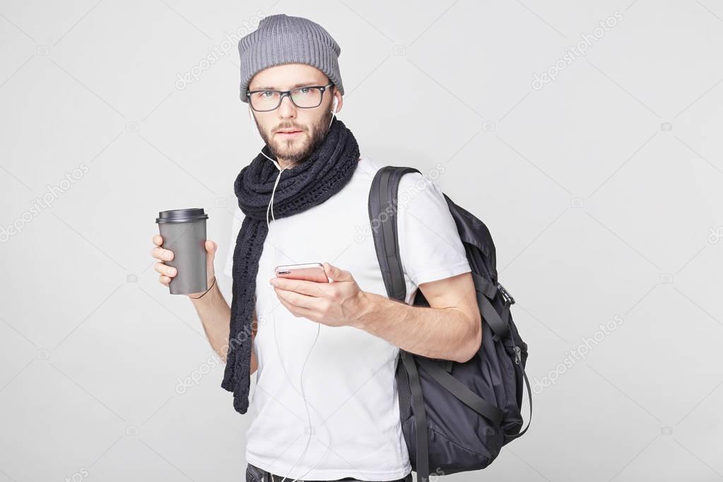 Serious bearded male traveler wearing spectacles, stylish hat and scarf, holding takeaway coffee in hand, looking with serious expression at the camera, holding phone, waiting for his companion call.