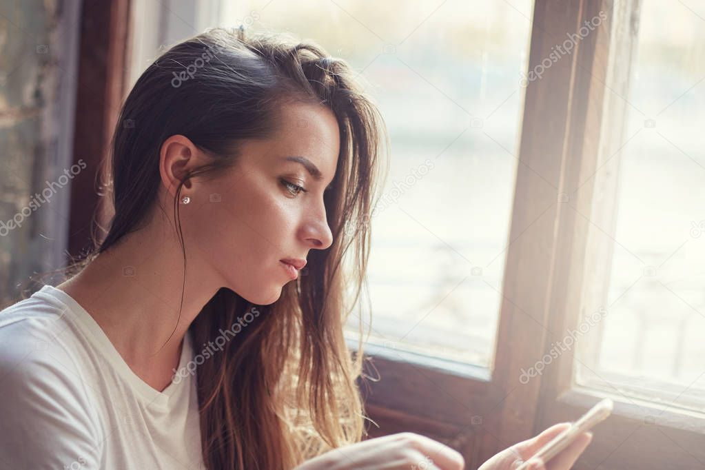 Close up shot of beautiful mixed race young brunette woman using mobile phone while sitting by window of modern cafe, texting her friend or lover, reading news with thoughtful expression on her face. 