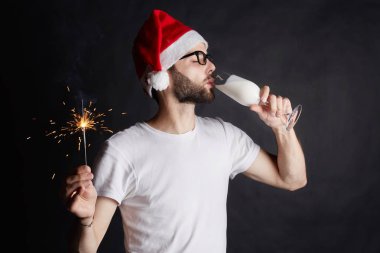 Portrait of handsome  man wearing Christmas hat with sparkler in hand during celebration catholic holiday. Young guy in stylish spectacles drinking yogurt at non-alcoholic party. Healthy lifestyle. clipart