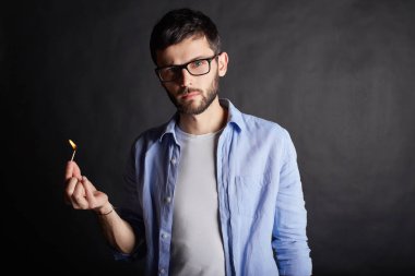 Elinde siyah stüdyo duvarda maç yanma ile çekici genç ciddi ya da tehlikeli beyaz bir adam portresi kapatın. Esmer hipster trendy gözlük kapalı elinde ateşle poz