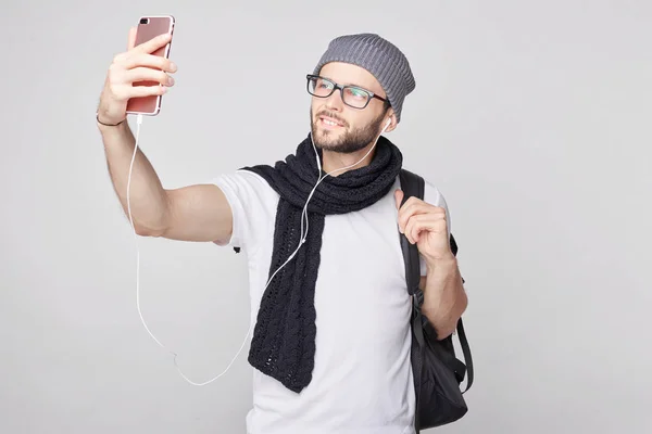 Guapo alegre joven hipster turista masculino con mochila celebración de teléfono celular para selfie. Joven viajero enérgico descansando por minuto después de largo viaje a pie, mirando felizmente en cámara de móvil . — Foto de Stock