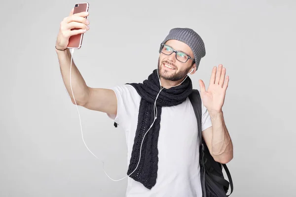 Personas, estilo de vida, viajes, turismo y tecnología moderna. Atractivo viajero caucásico en elegantes gafas y sombreros posando para selfie con sonrisa feliz y un gesto de hola contra la pared blanca . — Foto de Stock