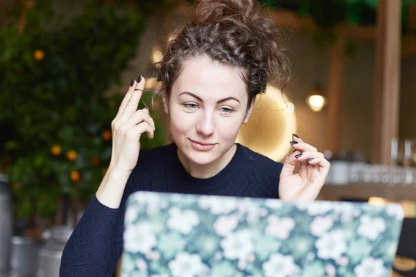 European woman keeps hands on curly locks, wants to have straight hair, in hurry doesn\'t know which hairstyle to do for special meeting at modern cafe. Lady uses monitor of laptop like a mirror.