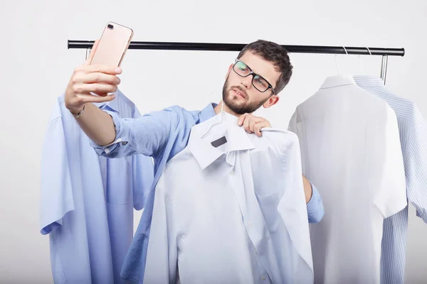 Metrosexual Caucasian guy taking self portrait on generic mobile phone, posing against blue background of his wardrobe, boasting about new stylish jeans and shirts she bought in the sale this morning.