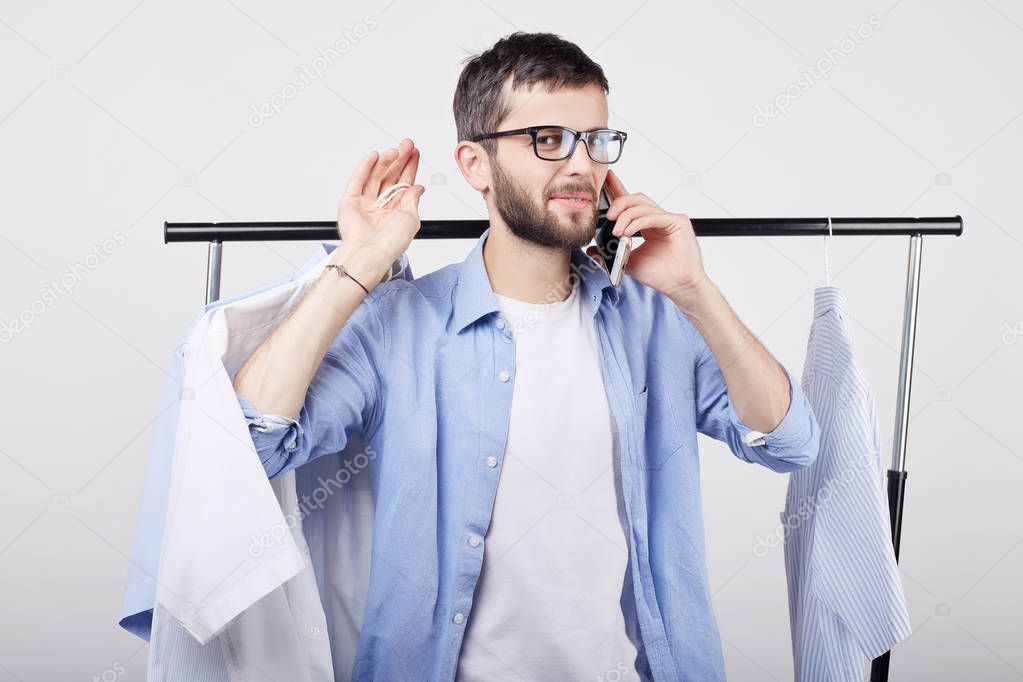 Attractive young Caucasian male shopaholic wearing stylish spectacles speaking on mobile phone with friend, boasting about purchases, shopping in city mall, standing at rack full of trendy clothes.