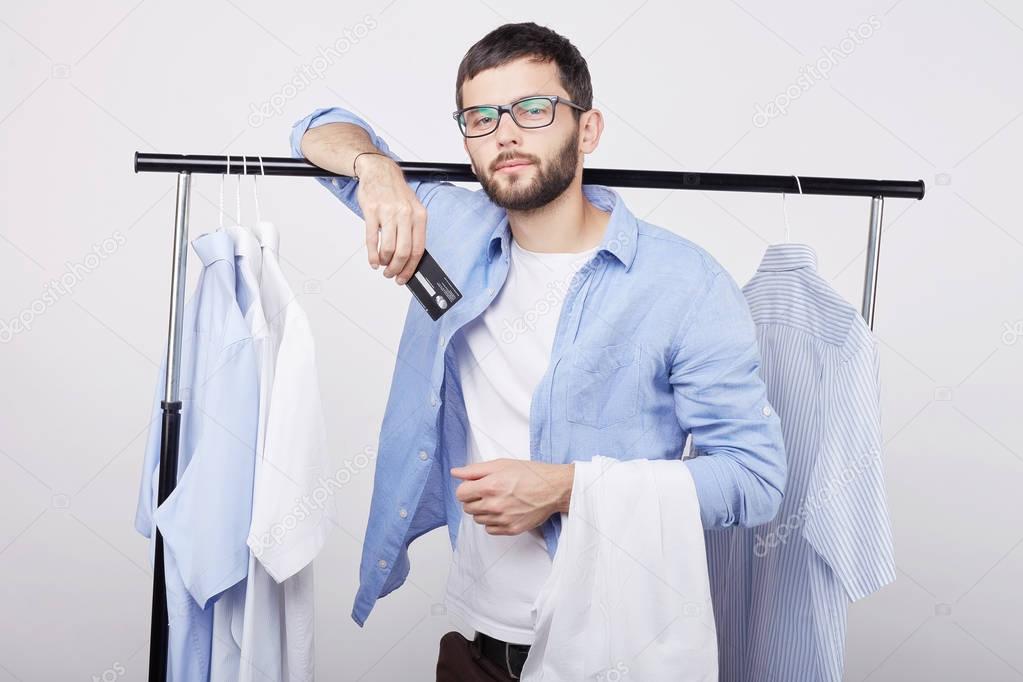 Shopping time. Smiling confident young Caucasian man, standing near hangers with clothes, demonstrating plastic card, being glad to receive salary going to buy new clothes in modern stylish showroom.