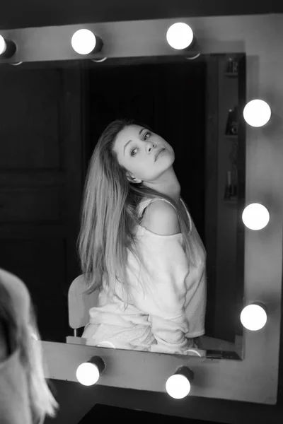 Vertical black & white portrait reflected young European seductive woman looking at mirror. Blond lady with long hair, admires herself, dressed casually, having serious mysterious face expression.