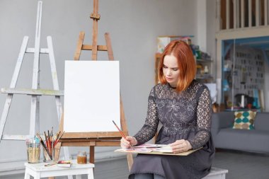 Güzel genç Kafkas kadın sanatçının Kızıl saçlı çizimler geniş atölyede ile lots-in boya iç yapım meşgul kapalı görünüm tablo ve sanat stüdyo alanı çevresinde mock up fırçalar.