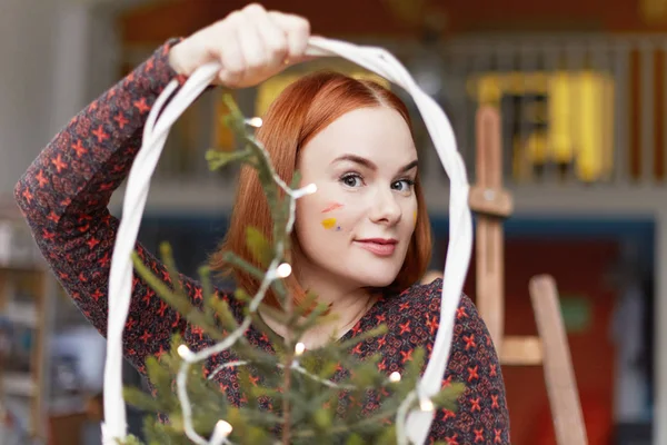 Portrait of creative female painter with oil colorful spots on face, ginger hair and freckles,holding small Christmas tree, looking at camera with serious expression, ready to celebrate New Year.