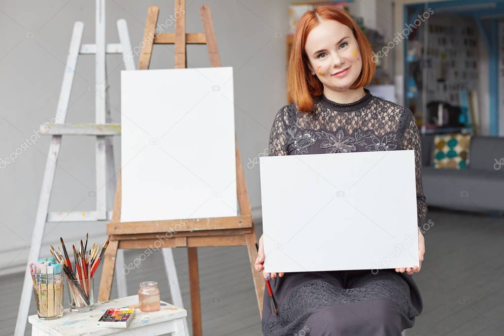 Cropped image of smiling Caucasian redhead hipster lady having an empty canvas mockup in hands with space for your advertising. Authentic lady sitting indoors in studio creating beautiful artworks.