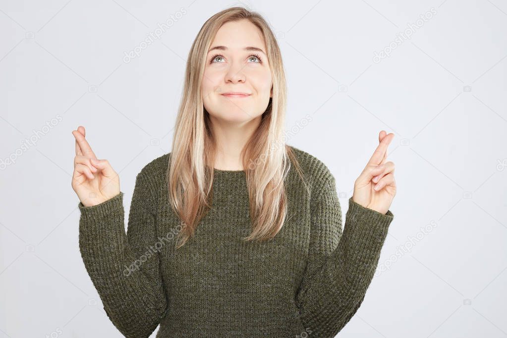 Young attractive female Caucasian student keeping her fingers crossed and eyes closed to make a desired wish. Charming white smile, positive heartwarming look of girl sincerely believing in victory.
