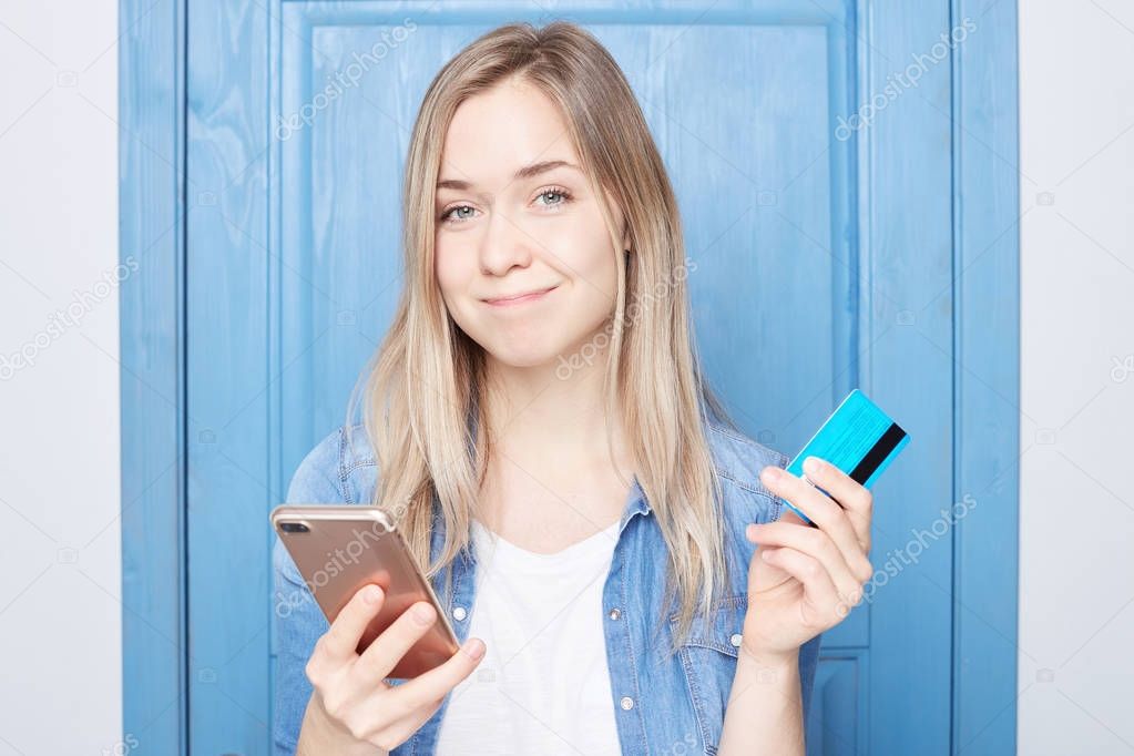 Indoor shot of beautiful woman with gentle smile, standing at home over blue door, being shopaholic, buying garment online, using smart phone and credit card. People, shopping, clothes concept.