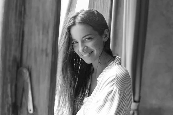 Indoor shot of amazing sophisticated young mixed raced woman wearing blue oversized shirt posing by window at light modern bedroom. Authentic lady looking at camera with cheerful pretty toothy smile.