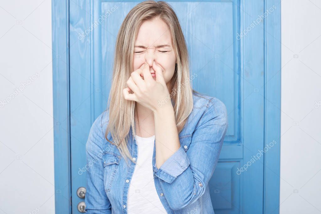 Displeased European cute female student with blond hair female plugs nose as smells something stink, unpleasant, feels aversion, isolated over blue door background. Young woman hates disgusting scent.