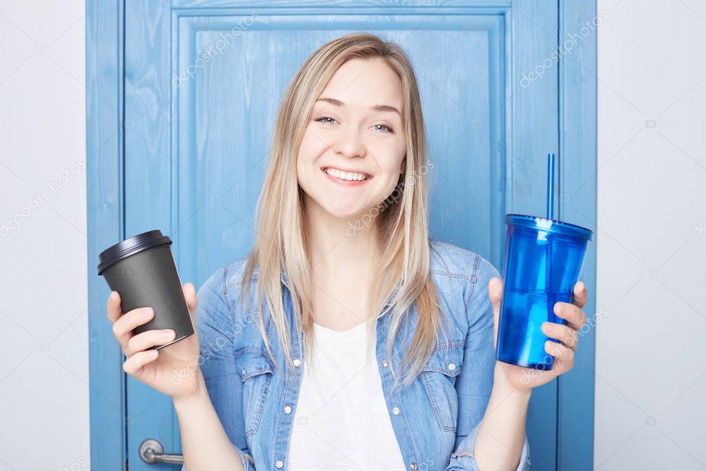 Beautiful young Caucasian female with wide happy toothy smile made choice between coffee and healthy beverage. Good looking blond American lady dressed in blue denim holds take away drinks in hands.