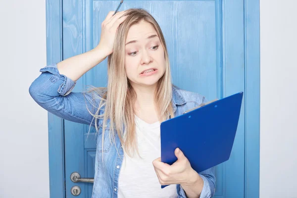 Failure, nervous breakdown, stress at work. Angry young European woman with notepad in hand grimacing and gesticulating, feeling stressed at office, desperate about problems, isolated on blue door.