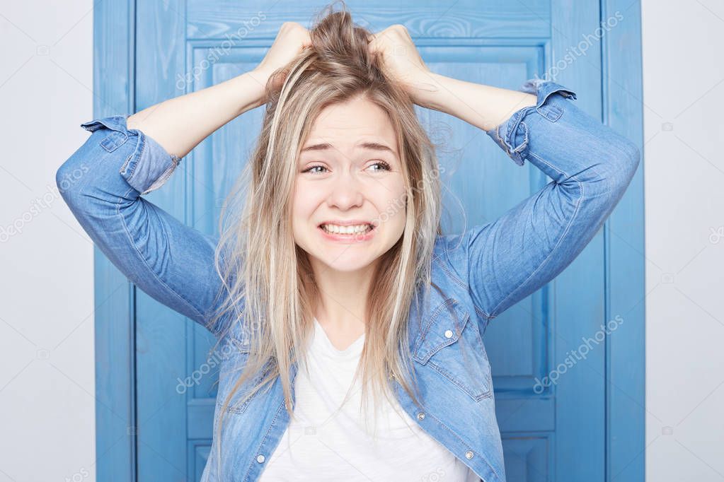 Isolated on blue door background young beautiful dressed casual Scandinavian woman is raging at herself, forgetting about important meeting, could not turn back time, grimacing and clowning from anger.