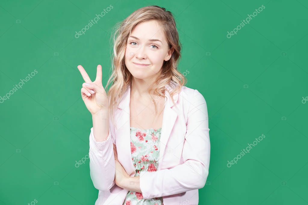 Fashionable female model with wavy hair, wears trendy colorful dress, round lips, gestures against green background, feels like star. Beautiful blonde woman shows peace sign. Body language concept.