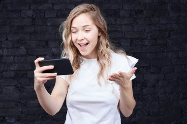 Sevimli genç sarışın Avrupa kadının samimi atış bağırarak ve heyecan içinde el hareketi siyah tuğla loft duvardan ayakta modern smartphone kullanarak rahat izlerken futbol maçı online, giyinmiş.