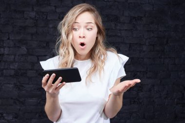 Sarışın genç kadın hoş görünümü ile akıllı telefon, dehşete ifade ile saatler haber tuğla arka plan üzerinde izole Web sayfasında, şok edici görünüyor. Kadın dijital telefon gösterir.