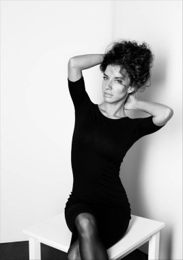 Siyah ve beyaz dikey stüdyo portre poz, kameraya bakarak ahşap sandalye üzerinde oturan çekici şık genç Kafkas kadın bir model. Kapalı otantik çekim kıvırcık saçlı Bayan davranıp elbise.