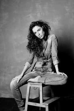 Siyah ve beyaz dikey studio kameraya bakarak büyük kulaklık kullanarak müzik ahşap sandalye üzerinde oturan çekici genç beyaz bir kadın portresi. Kot giyen Bayan otantik çekim.