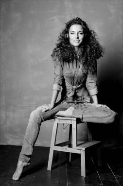 Siyah ve beyaz dikey studio kameraya gülümseyen büyük kulaklık kullanarak müzik ahşap sandalye üzerinde oturan çekici genç beyaz bir kadın portresi. Kot giyen Bayan otantik çekim. 