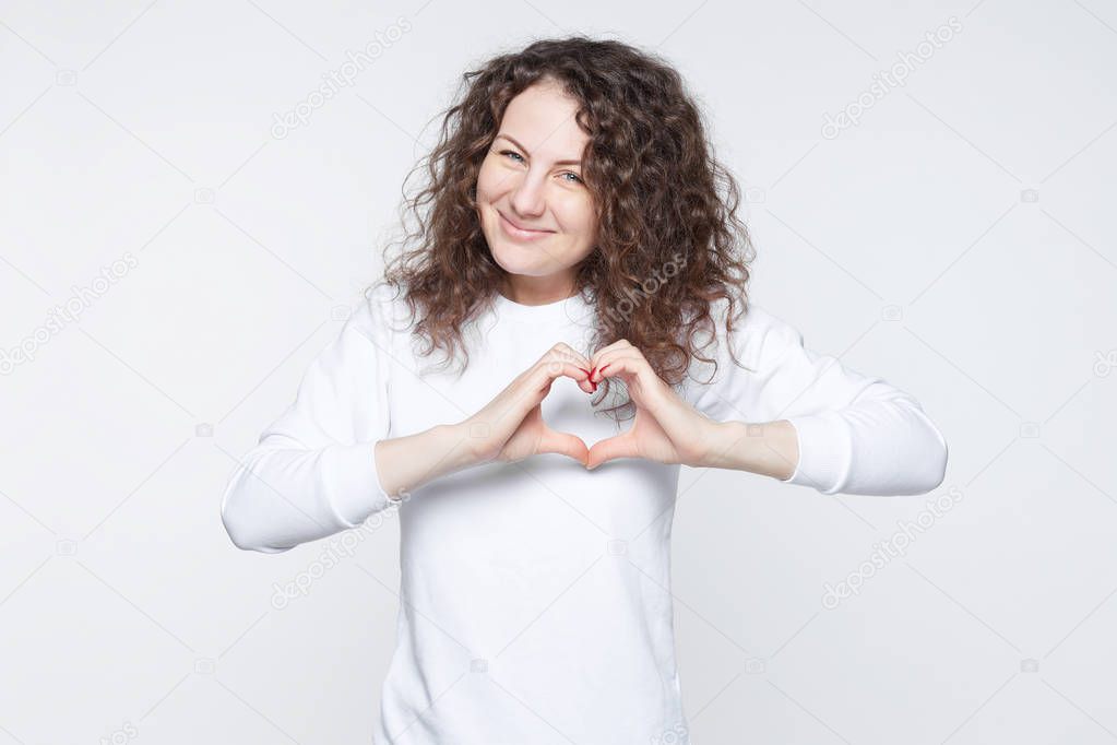 Indoor portrait of attractive American lady holding hands in shape of heart, symbolizing peace and unity. Blue eyed curly haired woman smiling tenderly at camera, showing love gesture on white wall.
