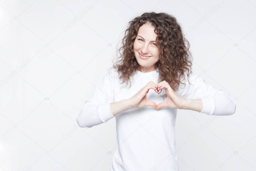 Studio shot of beautiful Caucasian lady holding hands in shape of heart, symbolizing love, peace and unity. Curly haired woman showing heart-shaped hand gesture, expressing affection and togetherness. 