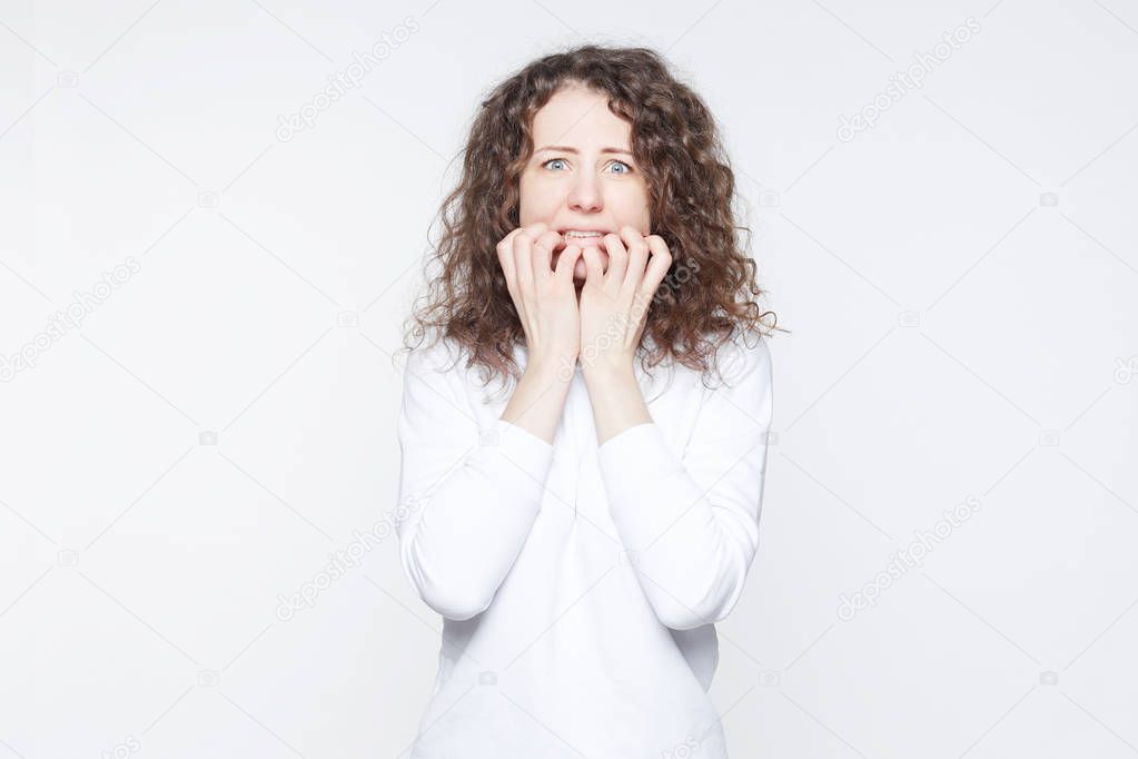Scared young European woman looks with blue bugged eyes at camera, being afraid of something, keeps hands on mouth, looks in terror, isolated over white background. Emotional horrified female indoor.