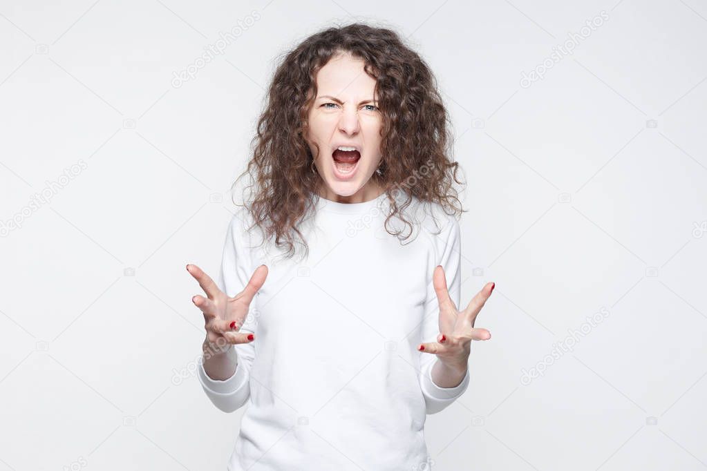 Close up isolated studio portrait of young annoyed angry woman holding hands in furious gesture. Young Caucasian female with bushy hair in white T-shirt. Negative human emotions, face expressions. 