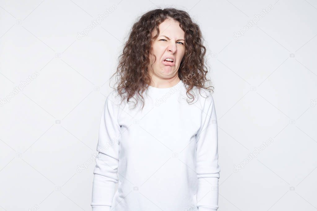 Stylish young female clenches teeth, has disgusting expression, refuses do something, makes grimace, isolated over white background. Dissatisfied youngster in casual white feels averse to do something