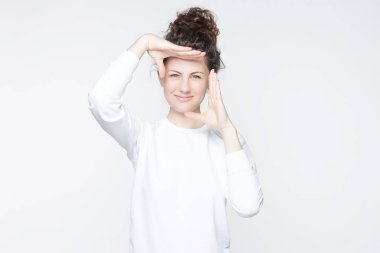 Genç çekici Avrupa Bayan kameraya gülümseyen ve izole beyaz duvar arka plan karşı dururken parmak çerçeve el hareketi kıvırcık saçları topuz ile kapalı stüdyo çekim. Beden dili kavramı.