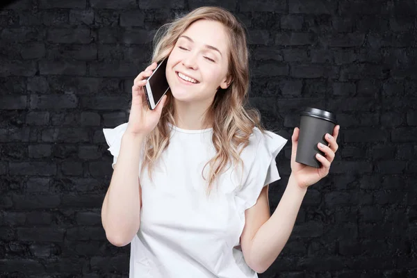 Happy Caucasian lady drinking coffee to go, talking on mobile phone with lover, closing eyes from pleasure to hear precious voice, while being away, posing against brick copy space wall for content.